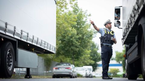 Uniformed police officer with a white cap on his head is standing on the road between two trucks. He is talking to the driver of the truck on the right and pointing to the left. There are parked cars in the background.